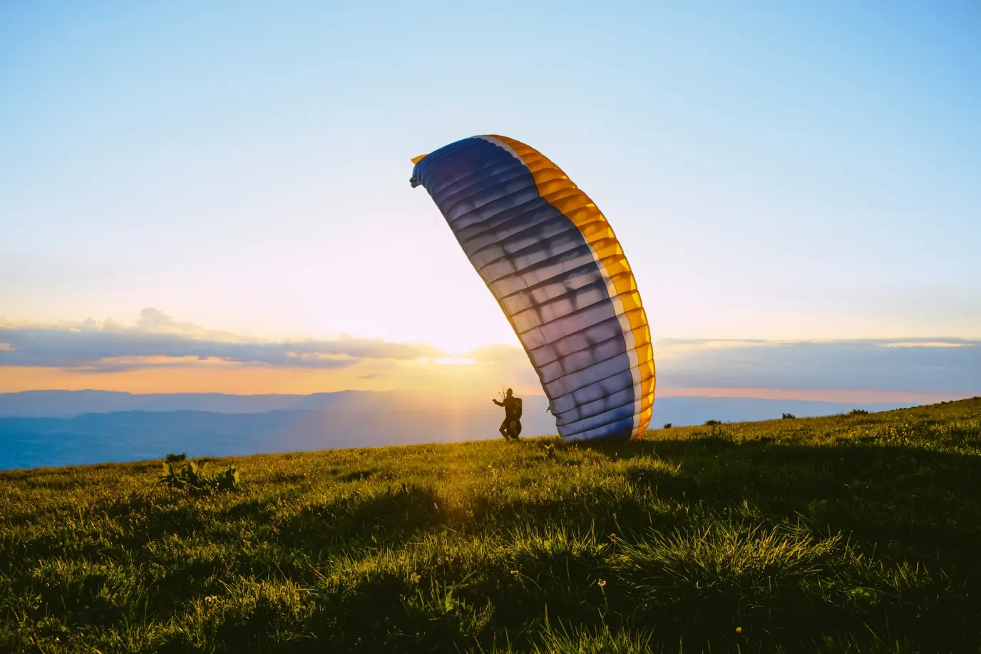 A paraglider on a grassy hilltop at sunset, the canopy inflated beside them.